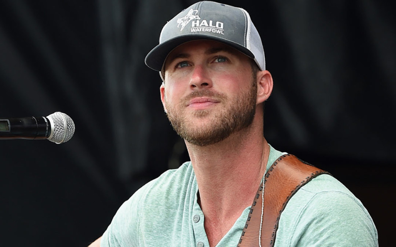 This image shows country singer Riley Green smiling while performing on stage, giving insight into his current relationship status as single.